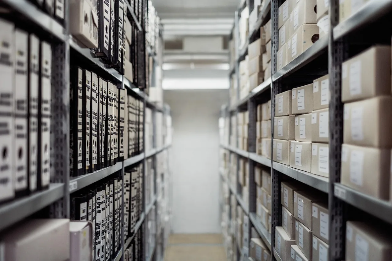 Pasillo entre dos estanterías metálicas llenas de archivadores y cajas de documentos etiquetadas