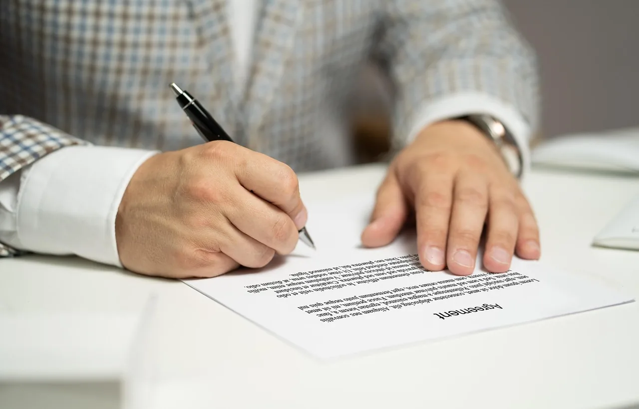 Hombre firmando un contrato impreso con bolígrafo sobre una mesa blanca, con la otra mano apoyada en el papel
